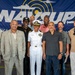 A group of approximately nine individuals pose for a formal group photo in front of a large blue 'NAVCUP' (Naval Cup) banner. One person in the center is wearing a white U.S. Navy dress uniform with officer insignia, medals, and a command cap, suggesting a military event or ceremony. The remaining individuals are dressed in civilian attire ranging from casual to business casual.
