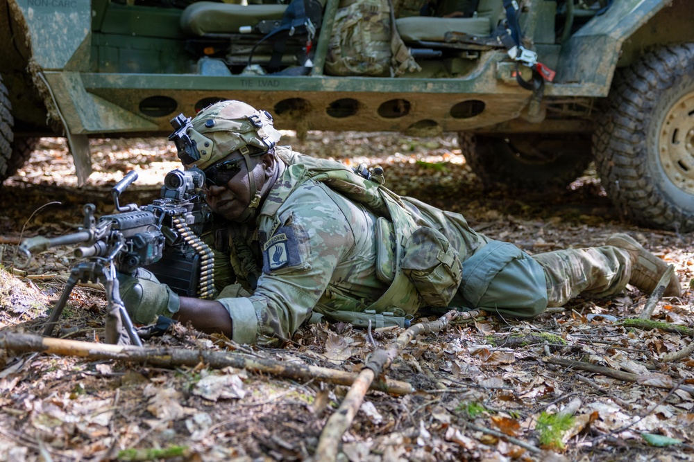 A soldier in full combat gear is lying prone on a forest floor in a firing position, aiming a belt-fed machine gun. The soldier is wearing MultiCam camouflage and displays a patch consistent with the 173rd Airborne Brigade on the left shoulder. A military tactical vehicle is visible in the background.