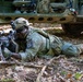 A soldier in full combat gear is lying prone on a forest floor in a firing position, aiming a belt-fed machine gun. The soldier is wearing MultiCam camouflage and displays a patch consistent with the 173rd Airborne Brigade on the left shoulder. A military tactical vehicle is visible in the background.