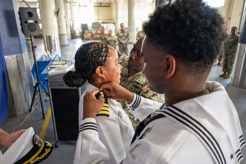 A U.S. Navy promotion ceremony is taking place in what appears to be a hangar or warehouse facility. A female naval officer is having insignia or collar devices pinned onto her white dress uniform by two individuals, consistent with a traditional Navy 'pinning' promotion ritual. Observers in Navy uniforms stand in the background watching the ceremony.