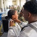 A U.S. Navy promotion ceremony is taking place in what appears to be a hangar or warehouse facility. A female naval officer is having insignia or collar devices pinned onto her white dress uniform by two individuals, consistent with a traditional Navy 'pinning' promotion ritual. Observers in Navy uniforms stand in the background watching the ceremony.
