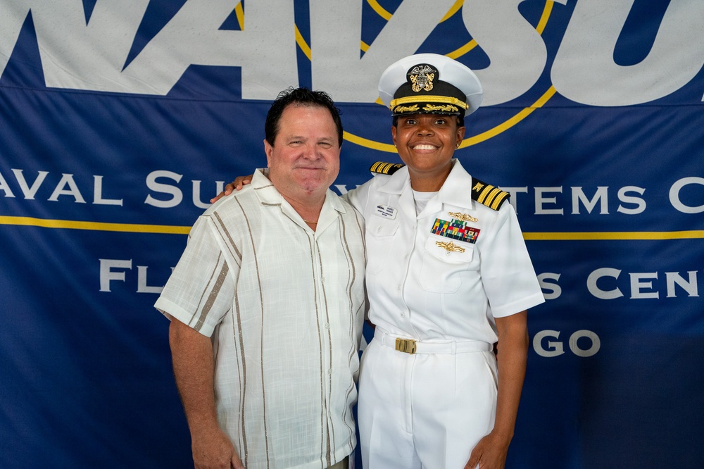 Two individuals pose together in front of a Naval Supply Systems Command banner, likely at a military ceremony or official event in San Diego. One person is in civilian attire while the other is a U.S. Navy officer in dress white uniform with commander-level insignia and multiple service ribbons. The setting and attire suggest a formal military recognition or change of command event.