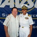 Two individuals pose together in front of a Naval Supply Systems Command banner, likely at a military ceremony or official event in San Diego. One person is in civilian attire while the other is a U.S. Navy officer in dress white uniform with commander-level insignia and multiple service ribbons. The setting and attire suggest a formal military recognition or change of command event.