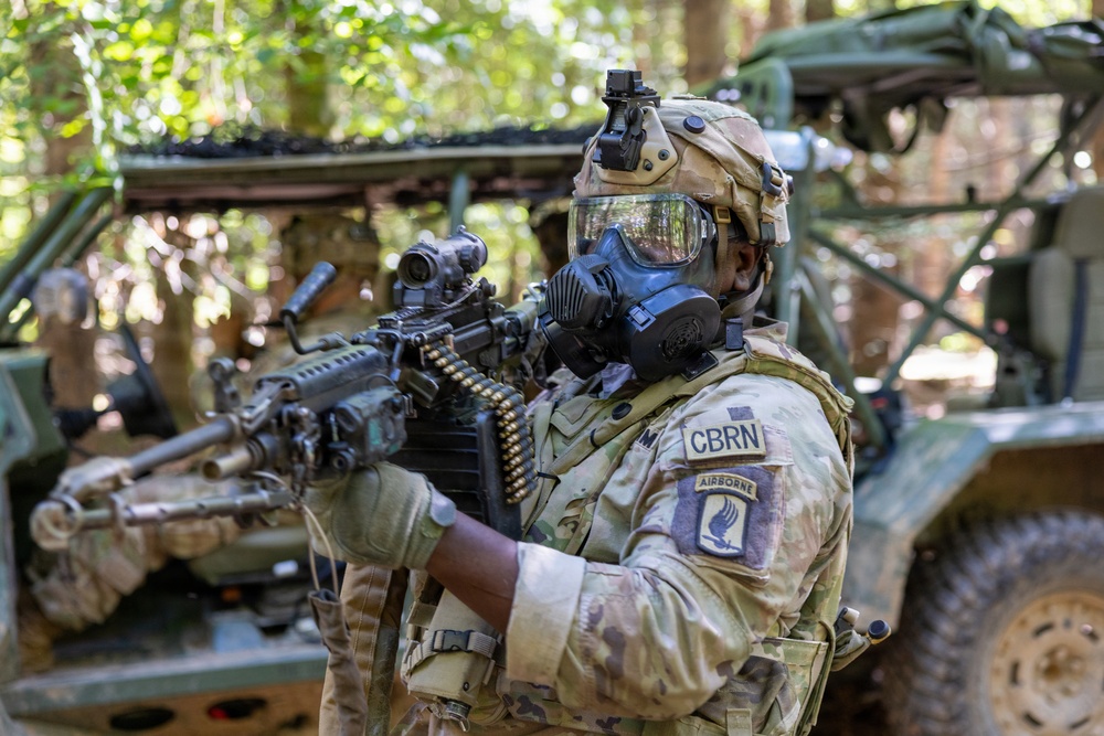 A U.S. Army soldier wearing full CBRN (Chemical, Biological, Radiological, and Nuclear) protective gear, including a gas mask and tactical helmet, is holding a belt-fed machine gun in a wooded environment. The soldier displays patches identifying them as CBRN-qualified and Airborne-qualified (13th Airborne Division or similar unit). Military vehicles are visible in the background, suggesting a field training exercise or operational scenario.