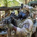 A U.S. Army soldier wearing full CBRN (Chemical, Biological, Radiological, and Nuclear) protective gear, including a gas mask and tactical helmet, is holding a belt-fed machine gun in a wooded environment. The soldier displays patches identifying them as CBRN-qualified and Airborne-qualified (13th Airborne Division or similar unit). Military vehicles are visible in the background, suggesting a field training exercise or operational scenario.