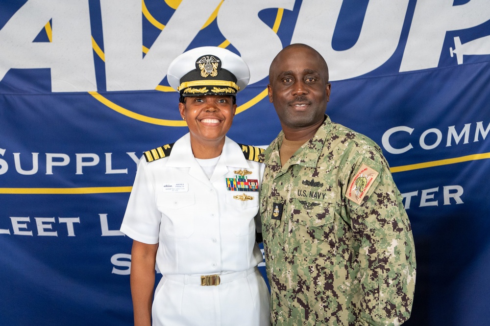 Two U.S. Navy personnel pose together in front of a NAVSUP (Naval Supply Systems Command) banner. The individual on the left wears white dress uniform with officer insignia, while the individual on the right wears Navy Working Uniform (NWU) camouflage. The setting appears to be an official military ceremony or event.