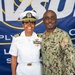 Two U.S. Navy personnel pose together in front of a NAVSUP (Naval Supply Systems Command) banner. The individual on the left wears white dress uniform with officer insignia, while the individual on the right wears Navy Working Uniform (NWU) camouflage. The setting appears to be an official military ceremony or event.