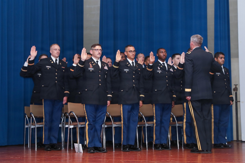 A group of U.S. Army personnel in dress blue uniforms are participating in a formal swearing-in or commissioning ceremony on a stage. They are raising their right hands while facing an officer who appears to be administering the oath. The ceremony is taking place in an auditorium or gymnasium with a blue curtain backdrop.