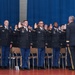 A group of U.S. Army personnel in dress blue uniforms are participating in a formal swearing-in or commissioning ceremony on a stage. They are raising their right hands while facing an officer who appears to be administering the oath. The ceremony is taking place in an auditorium or gymnasium with a blue curtain backdrop.