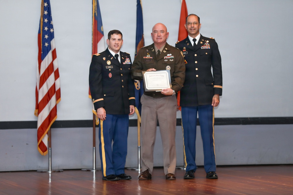 Three military personnel are posed together during what appears to be an awards or recognition ceremony. The central figure, wearing an olive/tan military uniform, is holding a framed certificate or award. Two flanking individuals are dressed in U.S. Army dress blue uniforms with visible rank insignia and ribbons.
