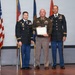 Three military personnel are posed together during what appears to be an awards or recognition ceremony. The central figure, wearing an olive/tan military uniform, is holding a framed certificate or award. Two flanking individuals are dressed in U.S. Army dress blue uniforms with visible rank insignia and ribbons.
