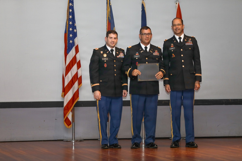 Three U.S. Army soldiers in dress uniforms are posed on a stage during what appears to be a formal military ceremony. The center soldier is holding a certificate or award folder, suggesting this is a recognition or awards ceremony. An American flag and additional military flags are visible in the background.