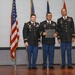 Three U.S. Army soldiers in dress uniforms are posed on a stage during what appears to be a formal military ceremony. The center soldier is holding a certificate or award folder, suggesting this is a recognition or awards ceremony. An American flag and additional military flags are visible in the background.