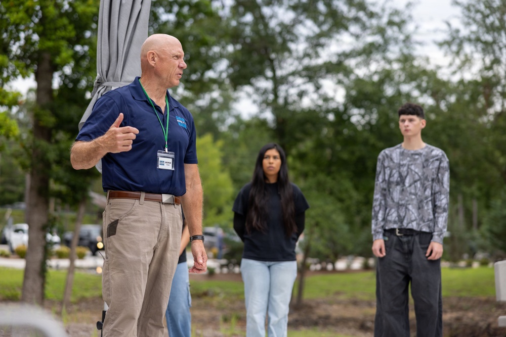 An older bald man wearing a navy polo shirt and a lanyard with a name badge appears to be speaking or presenting to a small group outdoors. Two younger individuals, a young woman in a black shirt and a young man in a gray patterned long-sleeve shirt, stand in the background listening attentively. The setting appears to be a landscaped outdoor area with trees and parked vehicles visible in the background.