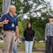 An older bald man wearing a navy polo shirt and a lanyard with a name badge appears to be speaking or presenting to a small group outdoors. Two younger individuals, a young woman in a black shirt and a young man in a gray patterned long-sleeve shirt, stand in the background listening attentively. The setting appears to be a landscaped outdoor area with trees and parked vehicles visible in the background.
