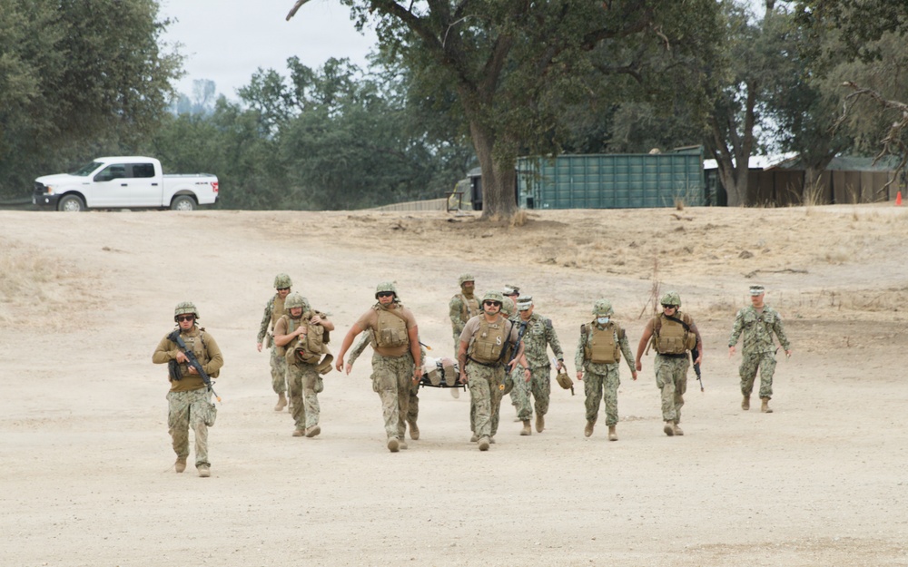 A group of military personnel in camouflage uniforms are conducting what appears to be a casualty evacuation (CASEVAC) training exercise on a dry, sandy terrain. Several personnel are carrying a litter (stretcher) with a simulated casualty while others provide armed escort. The setting includes trees, a green shipping container, and a white pickup truck in the background.