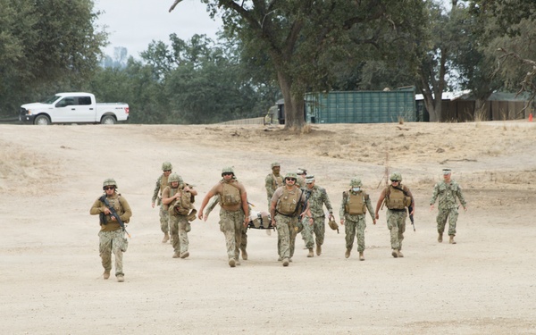 A group of military personnel in camouflage uniforms are conducting what appears to be a casualty evacuation (CASEVAC) training exercise on a dry, sandy terrain. Several personnel are carrying a litter (stretcher) with a simulated casualty while others provide armed escort. The setting includes trees, a green shipping container, and a white pickup truck in the background.