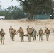 A group of military personnel in camouflage uniforms are conducting what appears to be a casualty evacuation (CASEVAC) training exercise on a dry, sandy terrain. Several personnel are carrying a litter (stretcher) with a simulated casualty while others provide armed escort. The setting includes trees, a green shipping container, and a white pickup truck in the background.