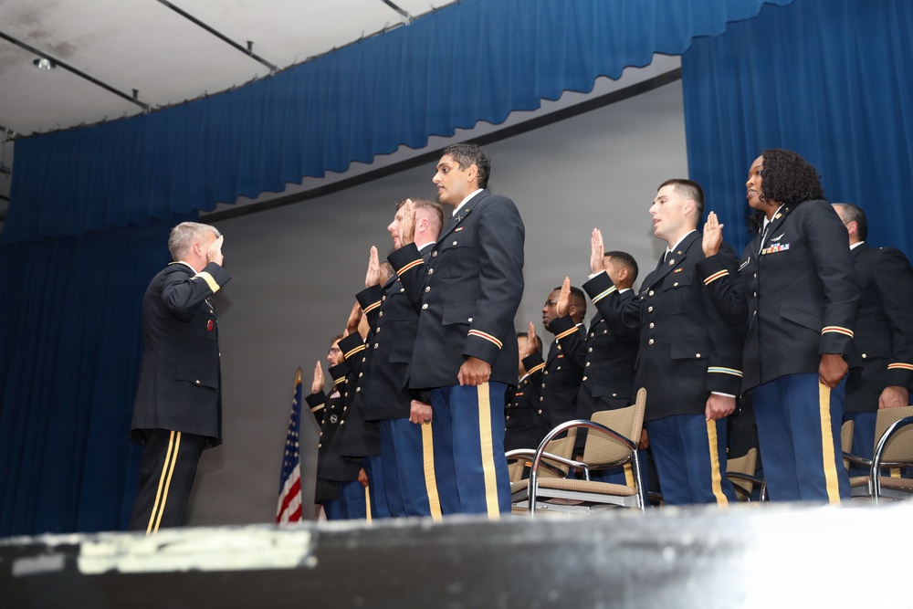 A military commissioning or oath-of-office ceremony is taking place on a stage. A senior officer on the left is administering an oath to a group of uniformed military personnel, who have their right hands raised. An American flag is visible in the background, and the setting appears to be an indoor auditorium or event hall with blue curtains.