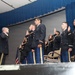 A military commissioning or oath-of-office ceremony is taking place on a stage. A senior officer on the left is administering an oath to a group of uniformed military personnel, who have their right hands raised. An American flag is visible in the background, and the setting appears to be an indoor auditorium or event hall with blue curtains.