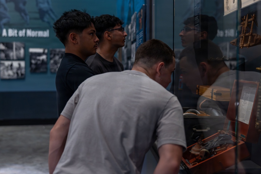 Three young men are examining museum display cases containing historical artifacts. The scene takes place in what appears to be a history museum, with wall-mounted photographs and text panels visible in the background. One visitor leans in closely to inspect items in a lower display case while the others view exhibits at eye level.