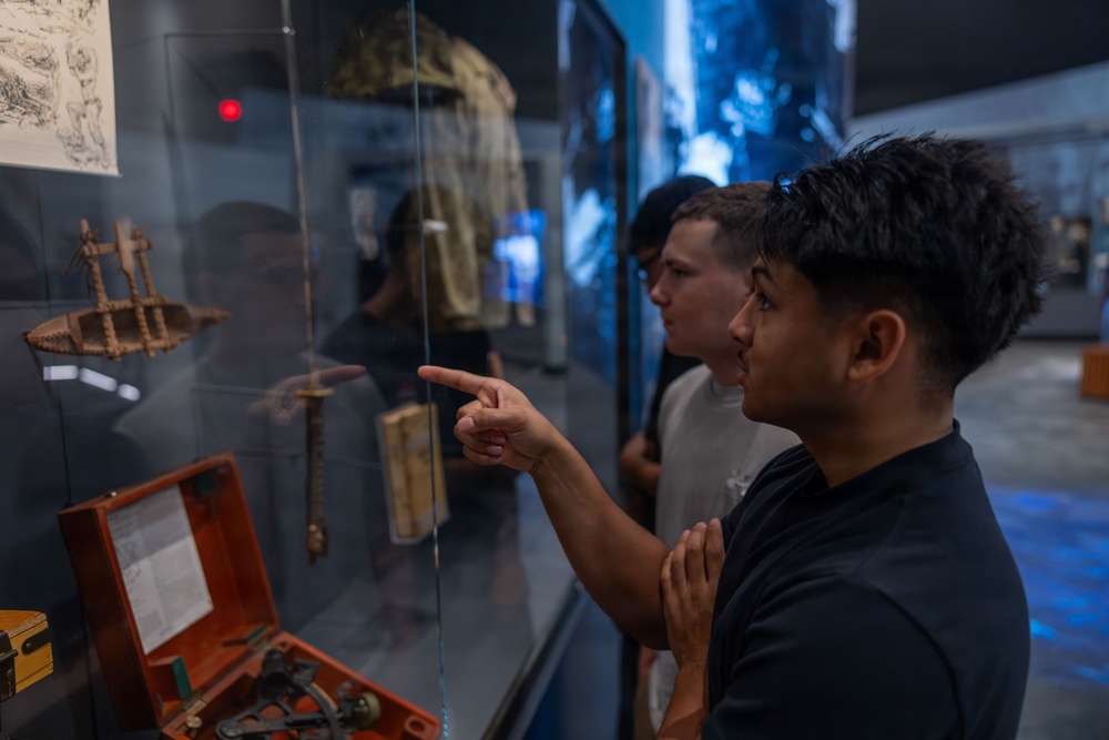 Two young men are examining artifacts inside a museum display case, with one pointing at an object of interest. The exhibit contains various historical items including what appears to be a wooden ship model, a dagger or blade, an orange case with tools, and military or period clothing in the background. The setting features blue ambient lighting typical of a modern museum environment.