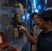 Two young men are examining artifacts inside a museum display case, with one pointing at an object of interest. The exhibit contains various historical items including what appears to be a wooden ship model, a dagger or blade, an orange case with tools, and military or period clothing in the background. The setting features blue ambient lighting typical of a modern museum environment.