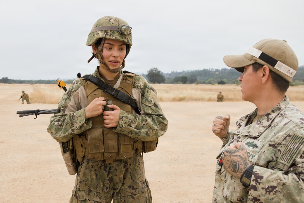 Two U.S. Navy personnel are engaged in conversation on an open training field. The individual on the left is in full combat gear including a helmet, body armor, and tactical vest, while the person on the right wears a patrol cap with a black band and Navy-labeled uniform. Additional personnel are visible in the background, suggesting a field training exercise.