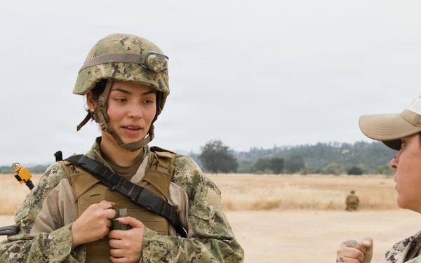 Two U.S. Navy personnel are engaged in conversation on an open training field. The individual on the left is in full combat gear including a helmet, body armor, and tactical vest, while the person on the right wears a patrol cap with a black band and Navy-labeled uniform. Additional personnel are visible in the background, suggesting a field training exercise.
