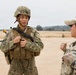 Two U.S. Navy personnel are engaged in conversation on an open training field. The individual on the left is in full combat gear including a helmet, body armor, and tactical vest, while the person on the right wears a patrol cap with a black band and Navy-labeled uniform. Additional personnel are visible in the background, suggesting a field training exercise.