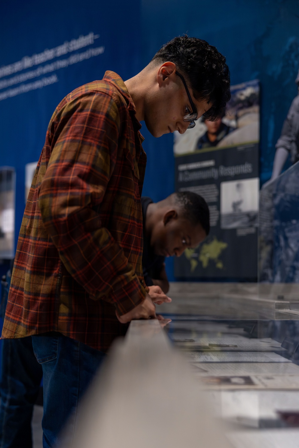 Two young visitors are leaning over a glass display case in what appears to be a museum exhibition. The foreground figure wears a red plaid flannel shirt and glasses, closely examining artifacts or documents inside the case. Behind them, exhibition panels with blue backgrounds display text and photographs, with one panel partially readable as 'A Community Responds.'
