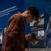 Two young visitors are leaning over a glass display case in what appears to be a museum exhibition. The foreground figure wears a red plaid flannel shirt and glasses, closely examining artifacts or documents inside the case. Behind them, exhibition panels with blue backgrounds display text and photographs, with one panel partially readable as 'A Community Responds.'