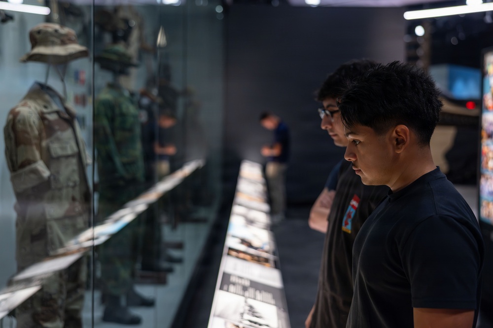 Two young visitors are examining a military uniform display case in what appears to be a military museum or exhibition hall. The display features multiple camouflage uniforms and equipment mounted behind glass, with an illuminated information panel running along the base. The scene captures an educational museum visit with visitors studying the exhibits closely.