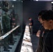 Two young visitors are examining a military uniform display case in what appears to be a military museum or exhibition hall. The display features multiple camouflage uniforms and equipment mounted behind glass, with an illuminated information panel running along the base. The scene captures an educational museum visit with visitors studying the exhibits closely.