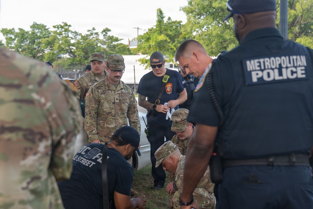 The image shows a multi-agency response scenario involving U.S. Army soldiers in OCP camouflage uniforms, Metropolitan Police officers, and what appear to be fire department personnel. Several individuals are crouched on the ground, suggesting a medical aid or casualty response situation. The gathering appears to be an inter-agency training exercise or an active emergency response.
