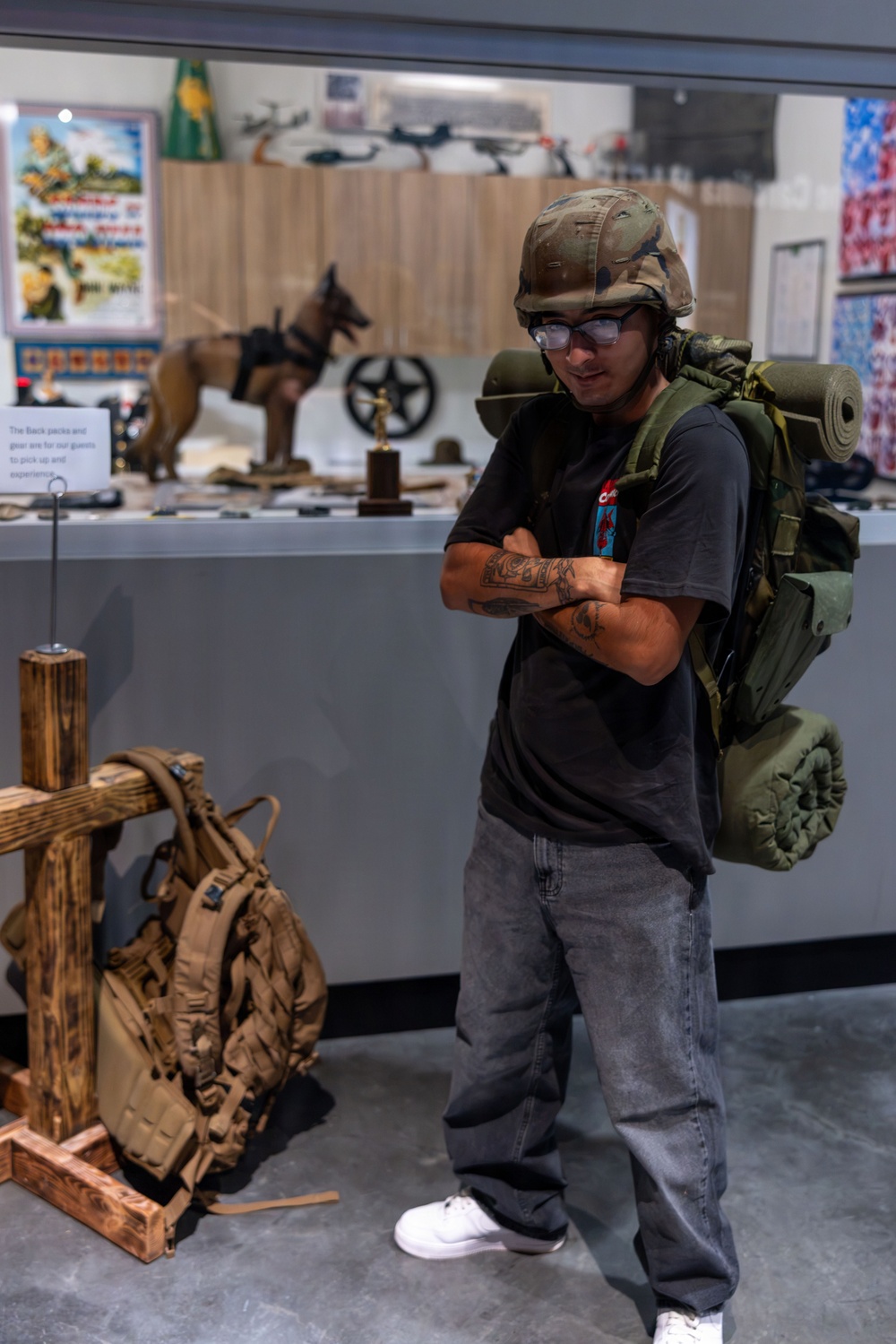 A young man wearing a military helmet and carrying a large olive-drab field pack with a sleeping roll stands in what appears to be a military museum exhibit. He is posing with arms crossed, wearing civilian clothes (black t-shirt, gray jeans, white sneakers) while trying on the display gear. A sign in the background indicates the backpacks and gear are available for guests to pick up and experience.