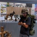 A young man wearing a military helmet and carrying a large olive-drab field pack with a sleeping roll stands in what appears to be a military museum exhibit. He is posing with arms crossed, wearing civilian clothes (black t-shirt, gray jeans, white sneakers) while trying on the display gear. A sign in the background indicates the backpacks and gear are available for guests to pick up and experience.