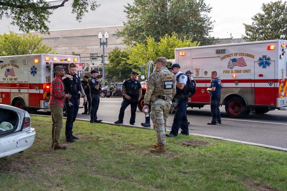 The image shows a multi-agency emergency response scene in Washington, D.C., with District of Columbia Fire &amp; EMS ambulances (units 19 and 3) deployed on a street. Personnel from multiple agencies are present, including DC Metropolitan Police officers, a National Guard soldier in camouflage uniform, and what appear to be plainclothes individuals. The scene suggests a coordinated response or staging operation rather than active medical treatment.