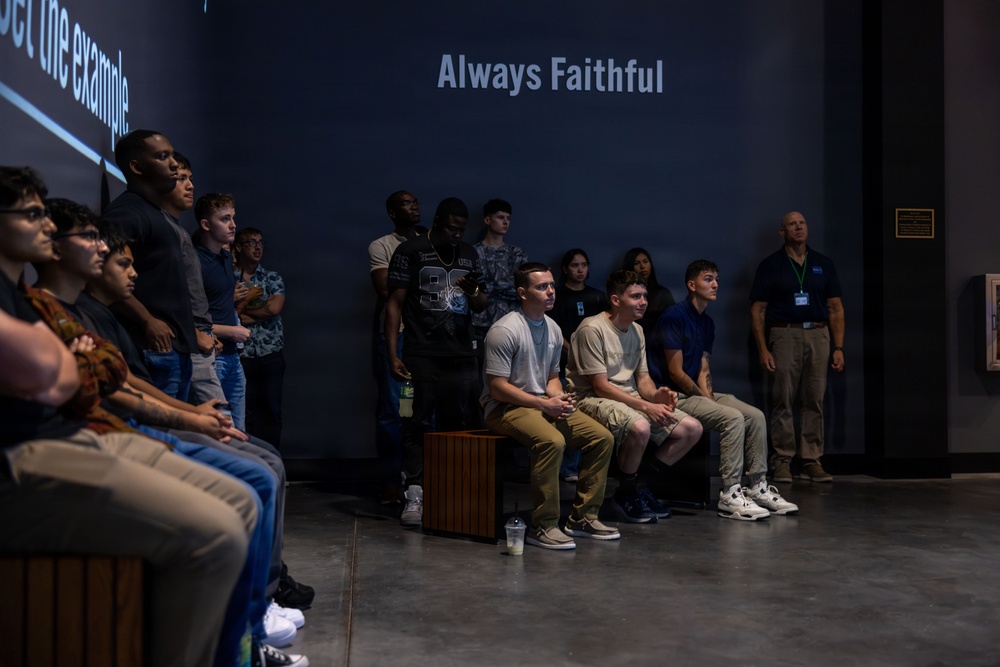 A group of young people are gathered in what appears to be a museum or exhibit hall, attentively watching or listening to a presentation. The dark-walled room features projected text reading 'Always Faithful' and partially visible text 'set the example' on the left wall. An older adult wearing a lanyard and badge stands to the right, likely serving as a guide or staff member.
