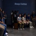 A group of young people are gathered in what appears to be a museum or exhibit hall, attentively watching or listening to a presentation. The dark-walled room features projected text reading 'Always Faithful' and partially visible text 'set the example' on the left wall. An older adult wearing a lanyard and badge stands to the right, likely serving as a guide or staff member.