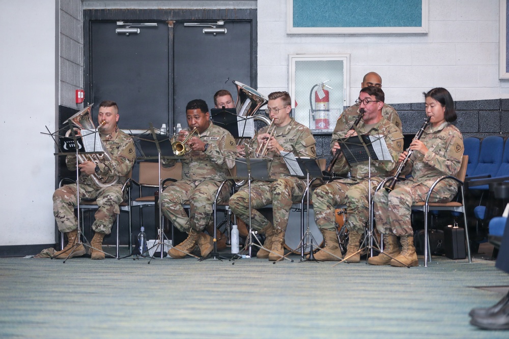 A group of U.S. Army soldiers in camouflage uniforms are performing as a military band ensemble in what appears to be an indoor facility. The musicians are seated in a row, playing various wind instruments while reading from music stands. The setting appears to be a formal or ceremonial performance event.