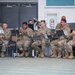 A group of U.S. Army soldiers in camouflage uniforms are performing as a military band ensemble in what appears to be an indoor facility. The musicians are seated in a row, playing various wind instruments while reading from music stands. The setting appears to be a formal or ceremonial performance event.