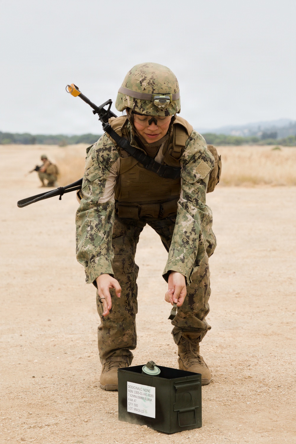 A military service member in digital camouflage uniform and combat gear is bending over an open ammunition can on a sandy training ground. The soldier is equipped with a rifle slung over their back, body armor, helmet with mounted light, and protective eyewear. A second soldier is visible in the background in a crouched position.