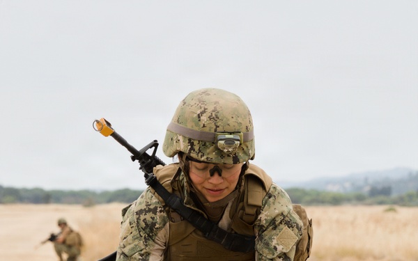 A military service member in digital camouflage uniform and combat gear is bending over an open ammunition can on a sandy training ground. The soldier is equipped with a rifle slung over their back, body armor, helmet with mounted light, and protective eyewear. A second soldier is visible in the background in a crouched position.