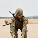 A military service member in digital camouflage uniform and combat gear is bending over an open ammunition can on a sandy training ground. The soldier is equipped with a rifle slung over their back, body armor, helmet with mounted light, and protective eyewear. A second soldier is visible in the background in a crouched position.