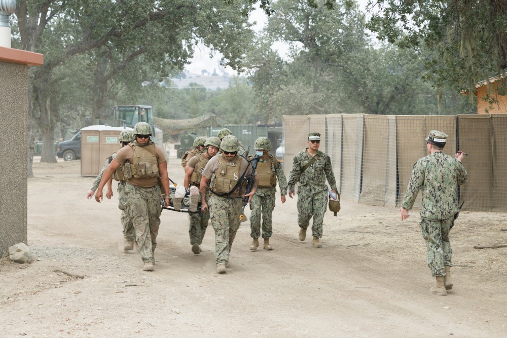 A group of military personnel in camouflage uniforms are carrying a casualty on a stretcher along a dusty dirt path in what appears to be a training exercise or field operation. Several personnel wearing tactical vests and helmets are transporting the litter while others walk alongside or ahead. The setting appears to be a military training facility with temporary structures, fencing, and oak trees in the background.