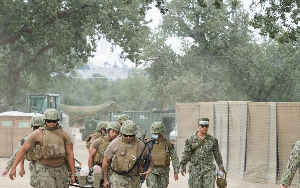 A group of military personnel in camouflage uniforms are carrying a casualty on a stretcher along a dusty dirt path in what appears to be a training exercise or field operation. Several personnel wearing tactical vests and helmets are transporting the litter while others walk alongside or ahead. The setting appears to be a military training facility with temporary structures, fencing, and oak trees in the background.