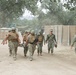A group of military personnel in camouflage uniforms are carrying a casualty on a stretcher along a dusty dirt path in what appears to be a training exercise or field operation. Several personnel wearing tactical vests and helmets are transporting the litter while others walk alongside or ahead. The setting appears to be a military training facility with temporary structures, fencing, and oak trees in the background.