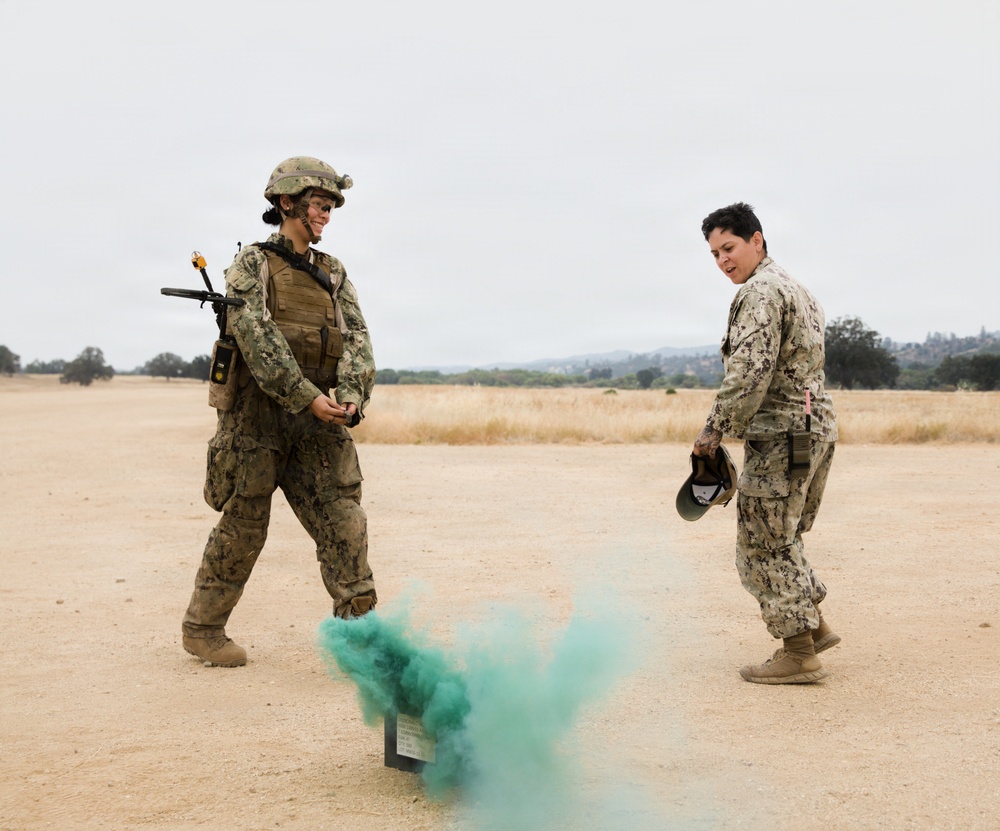 Two military personnel in camouflage uniforms are standing on an open dirt field observing a green smoke grenade or smoke signal device that has been activated on the ground between them. The person on the left is in full combat gear including a helmet, body armor, and a rifle, while the person on the right is in standard uniform holding a helmet. The setting appears to be a training exercise at a military installation.