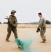 Two military personnel in camouflage uniforms are standing on an open dirt field observing a green smoke grenade or smoke signal device that has been activated on the ground between them. The person on the left is in full combat gear including a helmet, body armor, and a rifle, while the person on the right is in standard uniform holding a helmet. The setting appears to be a training exercise at a military installation.
